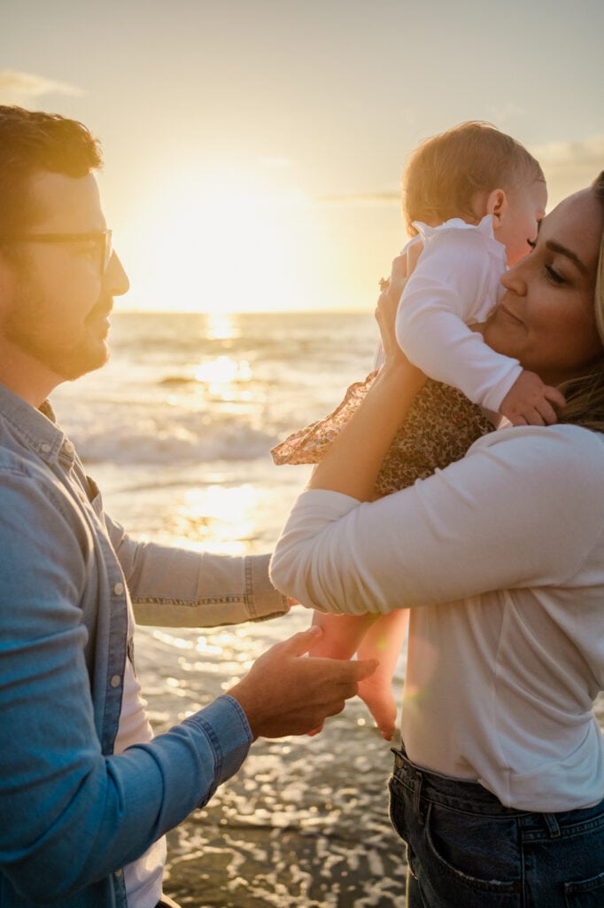 Dad hands mom baby daughter in front of setting sun over the ocean.