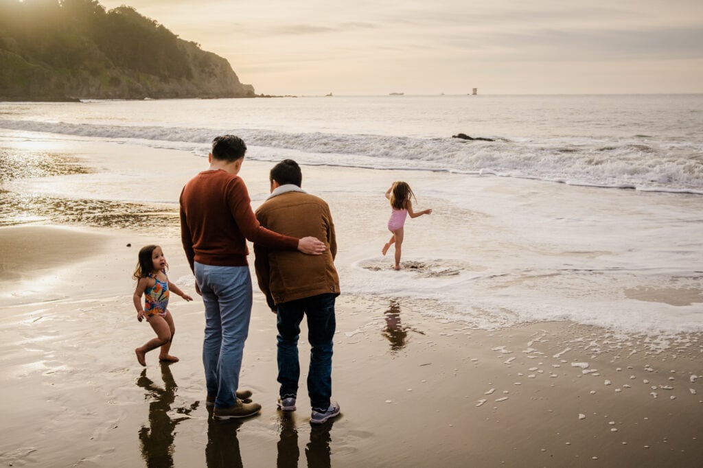 Two dads stand at the water's edge while their daughters play in the Pacific Ocean.