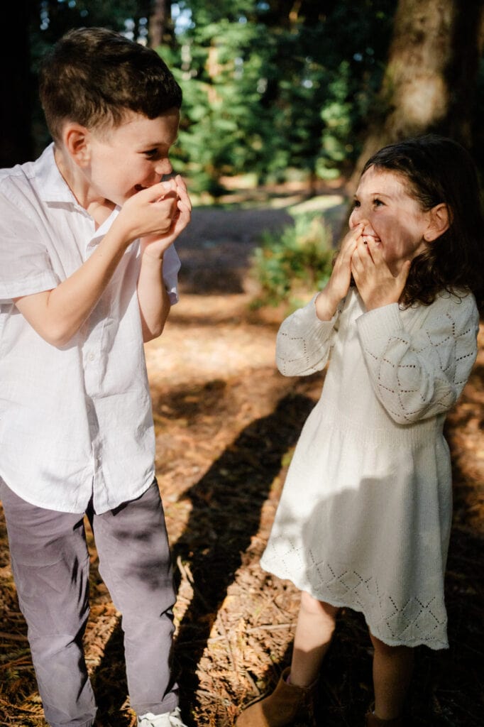 Two siblings cup hands over mouths while they look at each other and laugh.