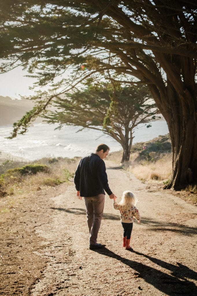 Dad holds daughter's hand as they walk down a road under a tree.