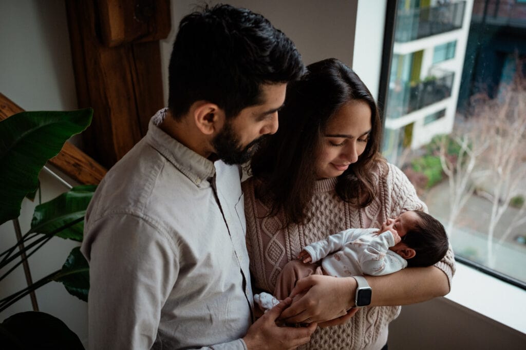 Mom holds newborn baby in front of window as dad stands next to her.