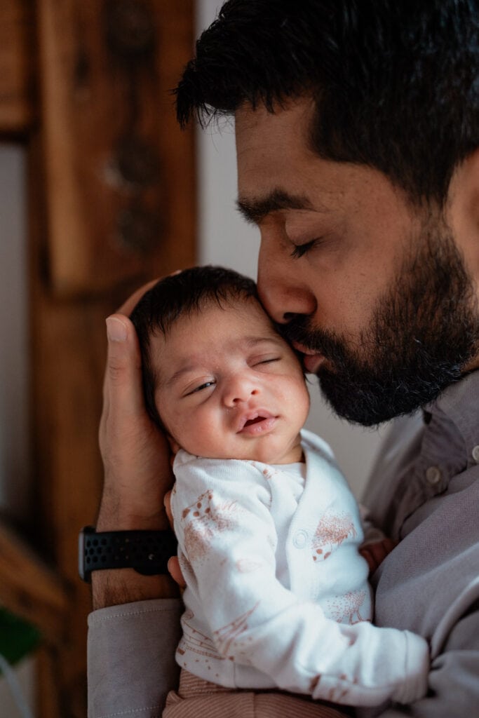 Dad kisses newborn with one eye barely open.