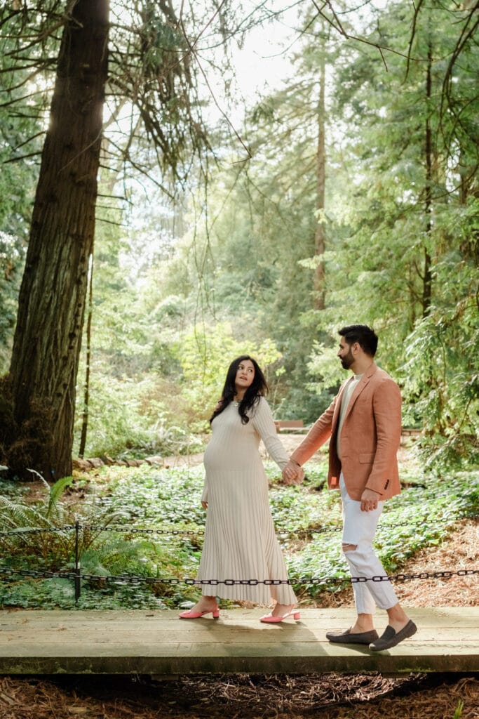 Pregnant couple walking hand in hand across footbridge beneath redwood trees.