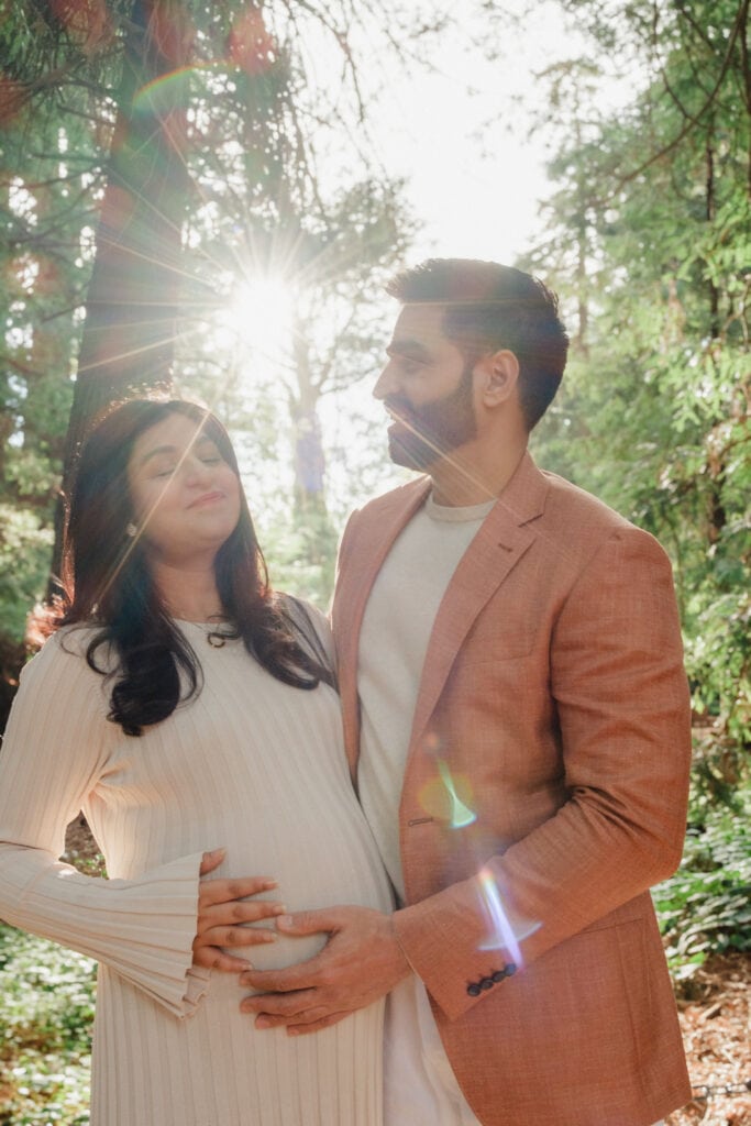 Man touches wife's pregnant belly as sun through the forest flares off of the camera lens.