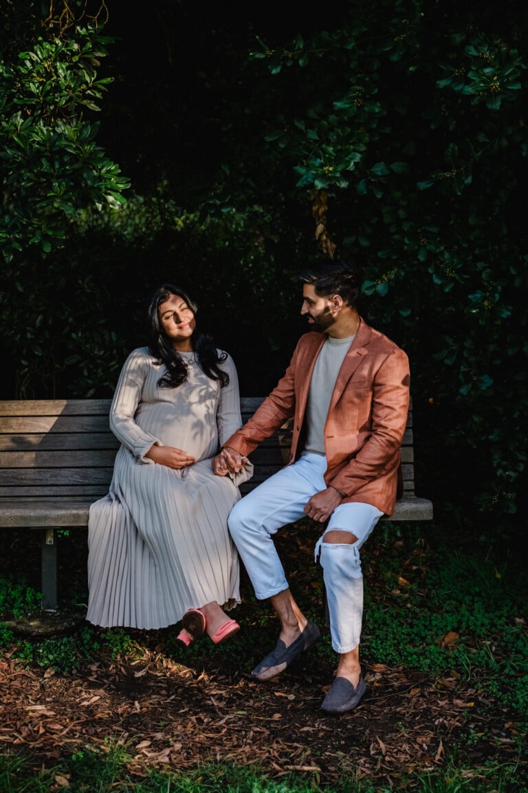Pregnant couple sit quietly on sun dappled park bench.