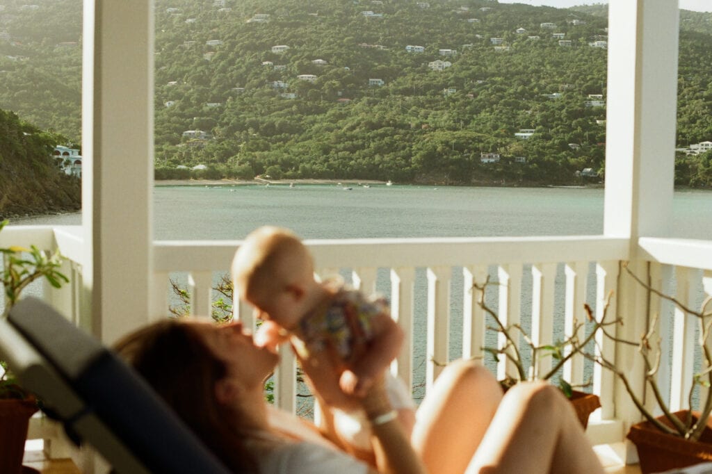 Woman holds baby above head in the foreground with a tropical beach behind.