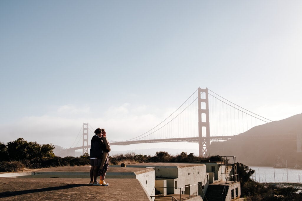Couple standing atop naval ruin in front of Golden Gate Bridge.