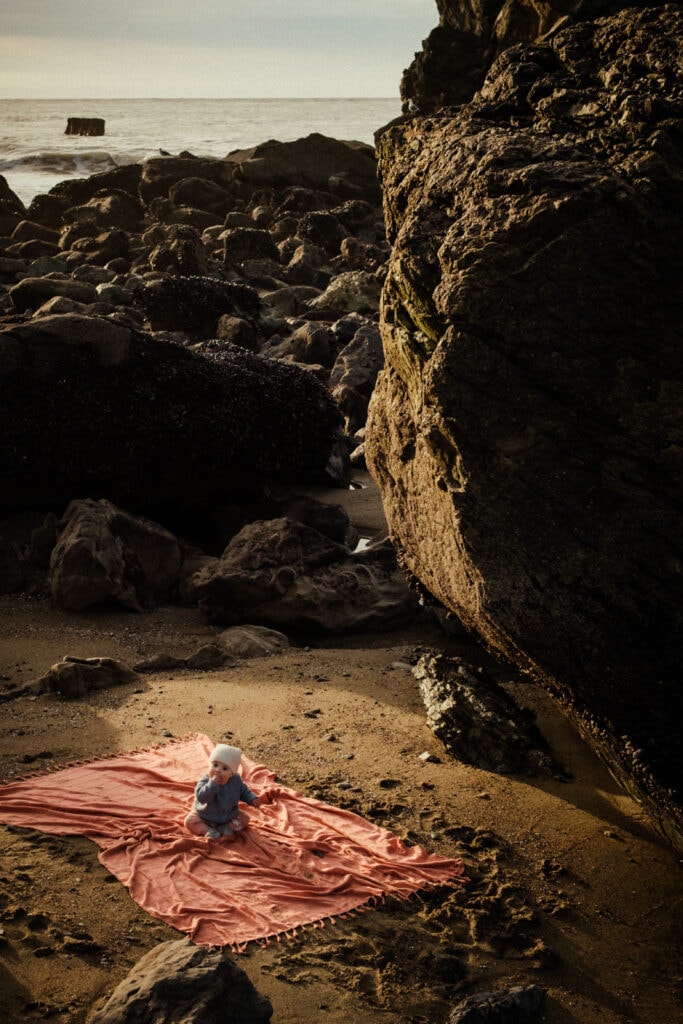 Baby sits on beach towel next to huge boulder in sunset light.