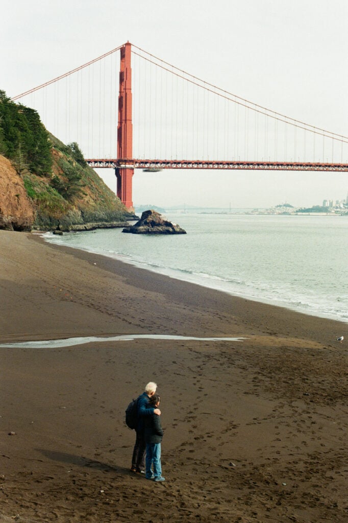 Couple stands on dark sand beach in front of Golden Gate Bridge.